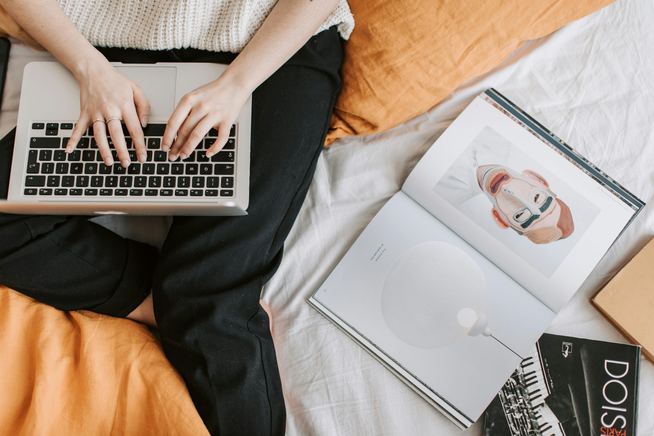 A woman in a cozy bedroom working on a laptop, surrounded by books and personal items.